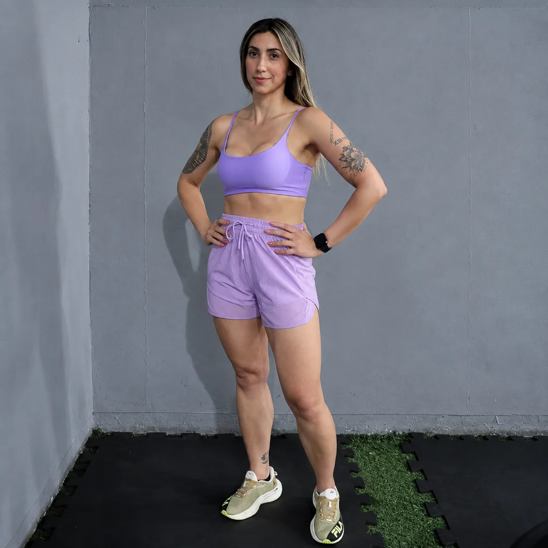 Modelo em top e short lilás, com a mão na cintura, em uma academia.